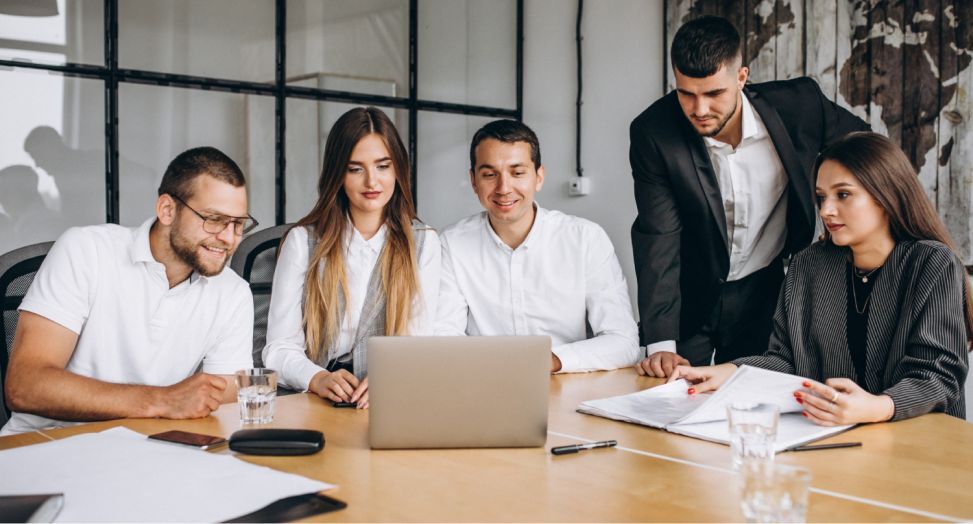 Five diverse professionals collaborate around a laptop during a business meeting in a modern office.